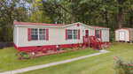 Single-story manufactured home with cream siding, red shutters, and a red roof. A red-railed porch leads to the front door. Neatly landscaped yard with flower beds and a small shed, bordered by trees in the background.