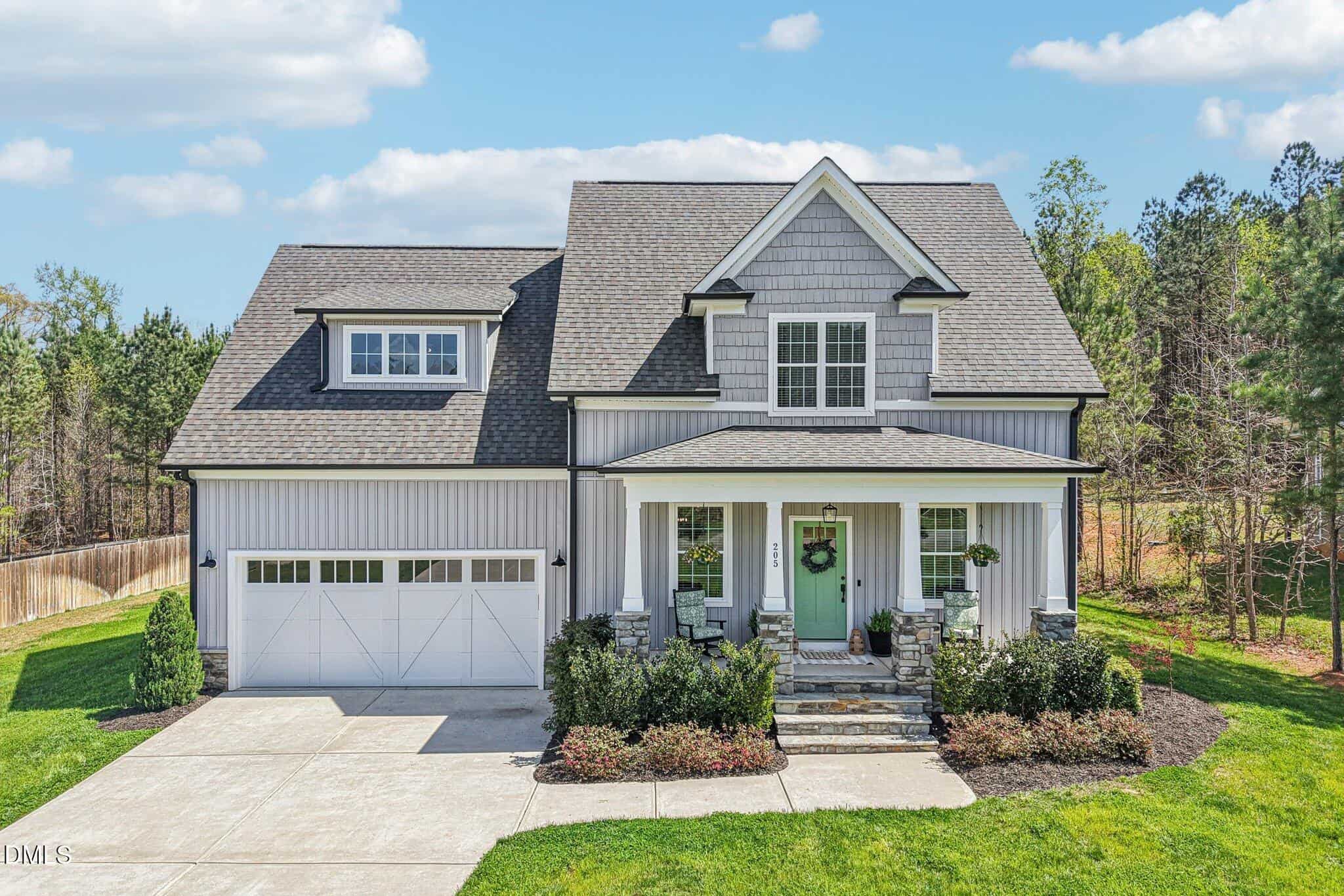 A modern two-story house with gray siding, a gable roof, double garage doors, and a covered front porch with stone columns, surrounded by green lawn, shrubs, and trees in the background.