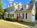 Two-story beige house with white trim, black shutters, dormer windows, and a covered front porch. Bushes and a small tree line the walkway; driveway and garage are visible to the right. Blue sky and trees in the background.