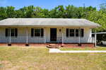 Single-story white house with black shutters and a covered front porch, surrounded by green trees. A sidewalk leads to steps up to the porch, and a small lawn is in front. A driveway and carport are visible to the right.