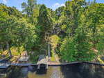 Aerial view of a lakeside property with a wooden dock and boathouse, lush green trees, terraced landscaping, and a house set back from the water among the trees.