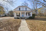 White two-story house with a blue front door, a covered porch, and two swings. Bare trees and autumn leaves cover the ground, and a curved walkway leads to the entrance. The sky is clear and blue.