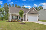 Single-story suburban house with beige siding, white trim, and a two-car garage. A small tree is planted in the front yard, and the lawn is freshly laid sod. Tall trees and a blue sky with clouds are in the background.