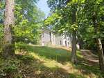 A light gray house with white trim sits on a grassy hill surrounded by tall trees and greenery, with a dirt driveway leading up to the home. Sunlight filters through the leaves, creating a peaceful, wooded setting.