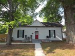 A small white house with black shutters, a red front door, a covered porch with two chairs and a table, and large trees in the front yard. A concrete walkway leads to the porch.