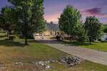 A single-story house with a front porch and two-car garage, surrounded by trees and a well-kept lawn, sits at the end of a long driveway at sunset with a colorful sky in the background.