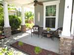 A cozy front porch with two wicker chairs, a small table with a flower vase, and a ceiling fan overhead. Pillows decorate the chairs, and there are plants and shrubs surrounding the porch.