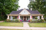 A charming light green house with a wide front porch, two American flags, potted plants, and a tidy lawn. Brick columns frame the porch, and large trees surround the home.