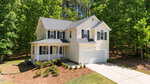 Two-story cream-colored house with dark gray roof, attached two-car garage, front porch, and driveway. The home is surrounded by green trees and has landscaped shrubs and mulch beds in front.