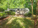 A beige and gray camper trailer is parked among green trees and bushes in a sunlit yard with grass and patches of shade. There are small steps leading up to the trailer entrance.