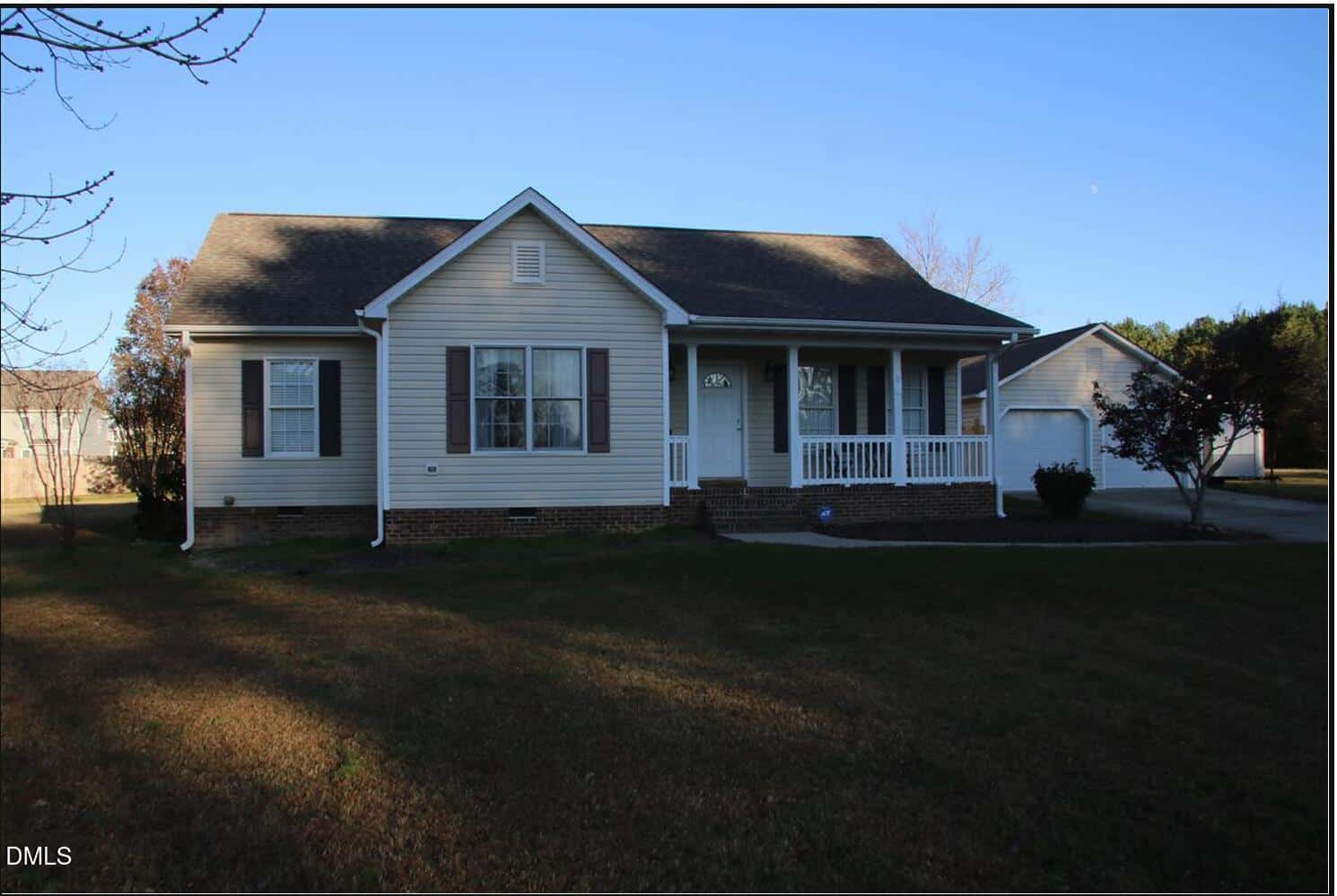A single-story beige house with white trim, brown shutters, and a covered front porch sits on a grassy lawn. A detached garage and driveway are visible to the right. Trees and clear sky are in the background.