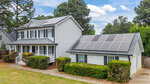 Two-story gray house with white trim and black shutters, featuring solar panels on the attached garage roof, surrounded by green bushes and trees under a partly cloudy sky.