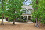 A two-story house with gray siding and white trim, a front porch with chairs, and surrounded by tall trees and a wooded yard. An American flag hangs near the porch, and a curved driveway leads to the home.