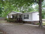 A white manufactured home with a brick foundation, covered front porch, and wooden railing. The house is surrounded by trees and has a gravel driveway in front.