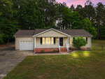 Single-story house with beige siding, a covered front porch, attached garage, and illuminated windows at dusk. Surrounded by grass lawn, mature trees, and a concrete driveway.