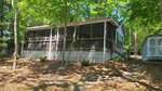 A single-story house with a large screened porch and lattice skirting, surrounded by trees. A gray shed sits to the right along a concrete path, and sunlight filters through the leafy canopy above.