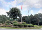 Landscaped entrance with a stone sign reading "Lake Royale," two flagpoles displaying American and North Carolina flags, a large tree, shrubs, and rocks, with woods and cloudy sky in the background.