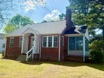 A small red brick house with a steep gray roof, white-trimmed windows, a screened porch on the right, a chimney, and a front door with a lattice panel. The house is surrounded by grass, trees, and shrubs.
