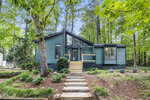 A modern blue house with large windows and a small front porch, surrounded by tall green trees and a landscaped yard with stone steps leading to the entrance.