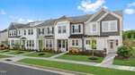 A row of modern two-story townhomes with white and gray exteriors, black roofs, and neatly landscaped lawns along a clean suburban street under a bright blue sky with scattered clouds.