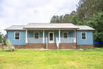 A single-story blue house with white trim and a metal roof, featuring a covered front porch with white railings and brick steps, set in a grassy yard with trees in the background.