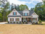 A modern farmhouse-style home with white siding, brown wooden shutters, a dark roof, a covered front porch, and landscaped shrubs, surrounded by trees and a partially grassed front yard.