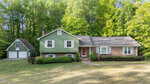 A split-level house with green siding and brick facade, white trim, and a shingled roof sits in front of a wooded area. A detached two-car garage with a basketball hoop is on the left. The lawn is spacious and well-kept.