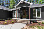 A modern dark gray house exterior with white trim, a covered front porch with wooden pillars, a wooden door with glass panels, and landscaped garden beds with small shrubs and mulch.