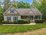 A beige two-story house with green shutters, a covered front porch, and an American flag. Well-maintained shrubs line the walkway, and tall trees are visible in the background.