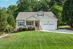 Single-story house with light siding, a covered front porch, and a two-car garage. The home is surrounded by well-maintained landscaping and green lawn, with trees in the background and a concrete driveway in front.