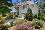 Two-story suburban house with stone and light-colored siding, arched windows, and a landscaped front yard featuring shrubs, flowers, and a small tree. A driveway leads to a single-car garage. Trees line the background.