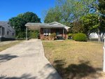 Single-story brick house with a small covered porch, driveway, and a sloped front yard with grass, bushes, and trees under a clear blue sky.