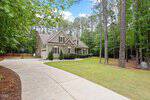 A beige house with white trim sits among tall trees. It has a manicured lawn, a curved driveway, and a basketball hoop near the garage. The house features a front porch and dormer windows under a blue sky.