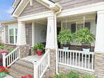 A charming front porch with stone columns, hanging ferns, two rocking chairs with blue pillows, a red potted plant, and a house number "123" on the pillar. Steps lead up to the porch, bordered by flowers and greenery.
