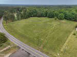 Aerial view of a large, grassy, open field bordered by trees, with a curved paved road running along one side and utility poles near the edge; the landscape extends into a forested area in the distance.