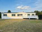 A single-story, light yellow manufactured home with green roof, white trim, and a large white front deck sits on a dry, grassy yard under a blue sky with a few clouds. Trees border the property.