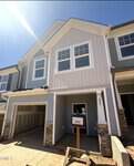 A newly constructed two-story townhouse with gray siding, white trim, a garage on the left, stone accents, and a signpost with construction information in front. Bright sunlight shines from the upper left corner.