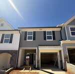 A newly constructed two-story townhouse with gray siding, white trim, black shutters, and an open garage under a clear blue sky. A temporary sign stands by the entrance, and the ground is bare dirt.