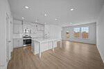 Modern, unfurnished kitchen and living space with white cabinets, stainless steel appliances, a center island with a sink, wood-style floors, large windows, and a door leading outside, all in a bright, open layout.