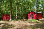 Two red wooden sheds stand among tall trees in a sunlit forest clearing. One shed is small with a window; the larger shed has double doors and a covered side area. The ground is covered with dirt and scattered leaves.