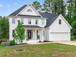 Two-story white brick house with black shutters, a front porch with rocking chairs, attached two-car garage, green lawn, small tree, and surrounded by tall trees under a blue sky.