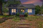 A small blue house with white trim and a dark roof, featuring a central green door, shuttered windows, and front bushes. The yard is patchy with grass, and the sky is colorful at sunset.