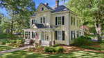 A large, two-story, light yellow Victorian-style house with white trim, a covered front porch, and an American flag, surrounded by green grass, trees, and shrubs on a sunny day.
