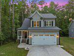 A two-story gray house with white trim, black shutters, and a front porch, surrounded by tall trees. The driveway leads to a double garage, and the sky is purple at sunset.