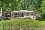 Single-story house with light brick exterior, a covered carport on the left, decorative white trim, landscaped front yard, and surrounded by tall trees in a wooded area.