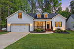 A modern white house with black trim at 1427 Sagamore Drive, Louisburg features a double garage, front porch with chairs, landscaped yard, and warm lights glowing at dusk. Tall trees and neighboring houses complete the inviting scene.