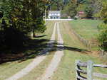 A long, narrow gravel driveway curves through a grassy field lined with wooden fences, leading to a white two-story house surrounded by trees under a clear blue sky.