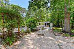 At 120 Pawnee Drive, Louisburg, a small white house with a screened porch sits among tall green trees. Its yard features gravel paths, landscaped beds, and a wooden trellis covered with climbing plants as sunlight filters through the leaves.