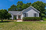 A single-story white house with gray roof at 200 Bradford Ridge Drive, Youngsville, is surrounded by green trees and bushes on a lawn with patches of dry grass under a clear blue sky.