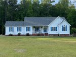 A single-story white house with blue shutters and a gray roof sits on a large grassy lawn at 113 Mayfield Place in Youngsville, surrounded by tall green trees. There is a small front porch with white columns and hanging plants.
